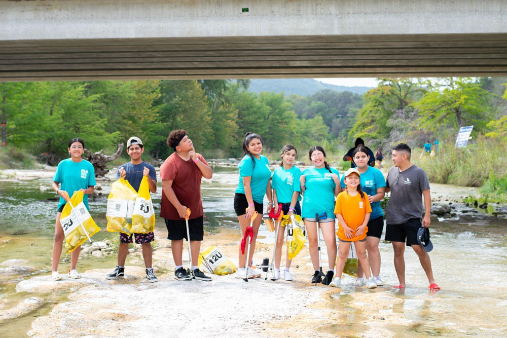 A group of people wearing matching blue shirts stand together under a bridge, holding yellow bags, likely participating in a community clean-up event by a river in Uvalde County, Texas