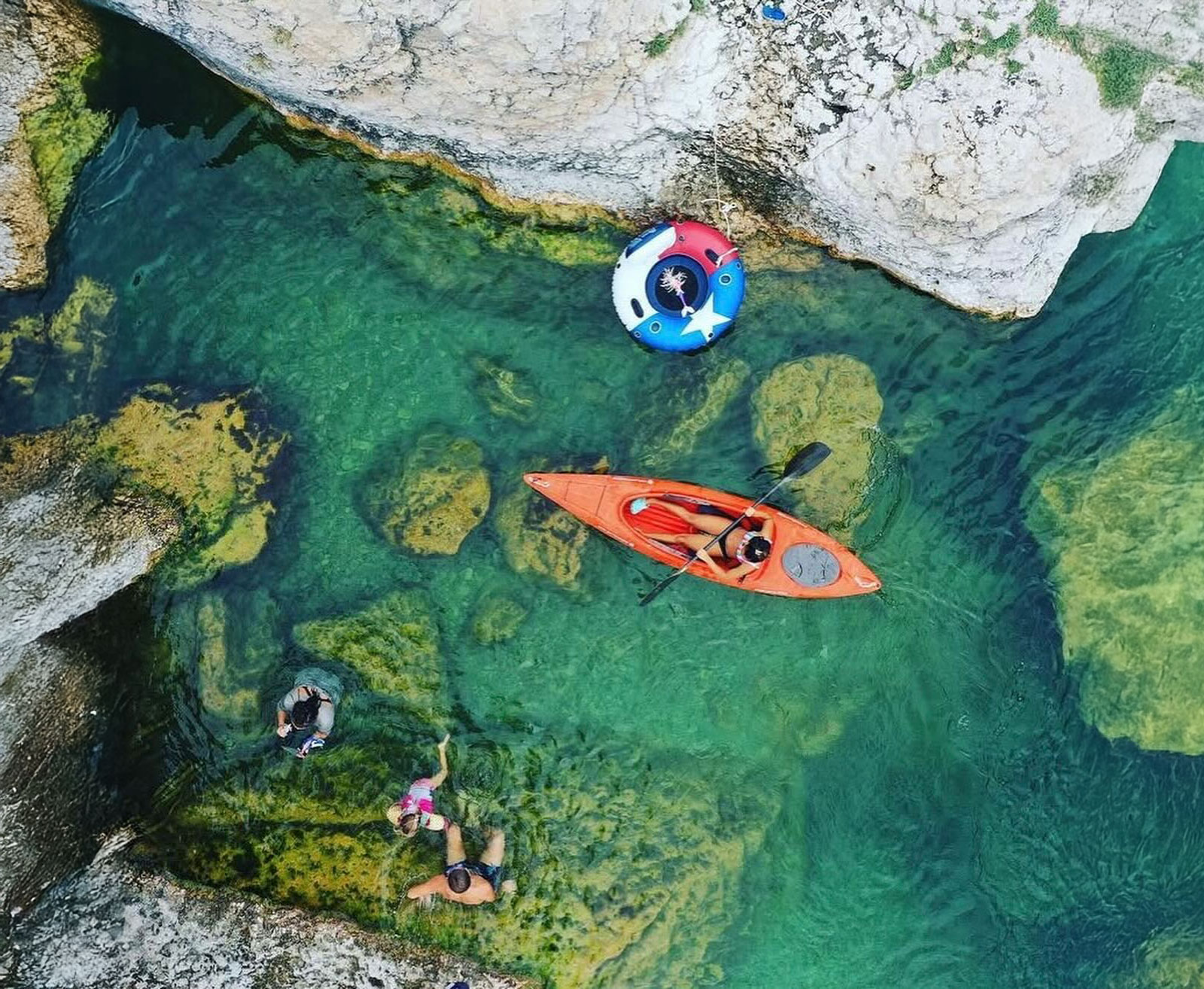 Aerial view of a serene river with kayakers, swimmers, and colorful floating devices amidst clear, turquoise waters and rocky outcrops in Uvalde County, Texas