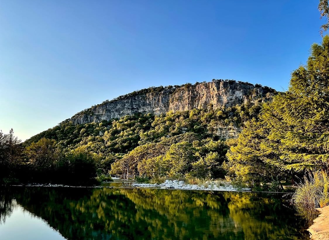 A serene landscape featuring a calm lake reflecting the surrounding trees and a rocky mountain under a clear blue sky in Uvalde County, Texas