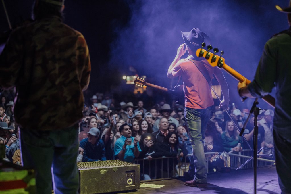 Live Music 3 A musician in a red shirt and cowboy hat plays an electric guitar on stage, backed by a large, enthusiastic crowd at a nighttime outdoor concert in Uvalde County, Texas