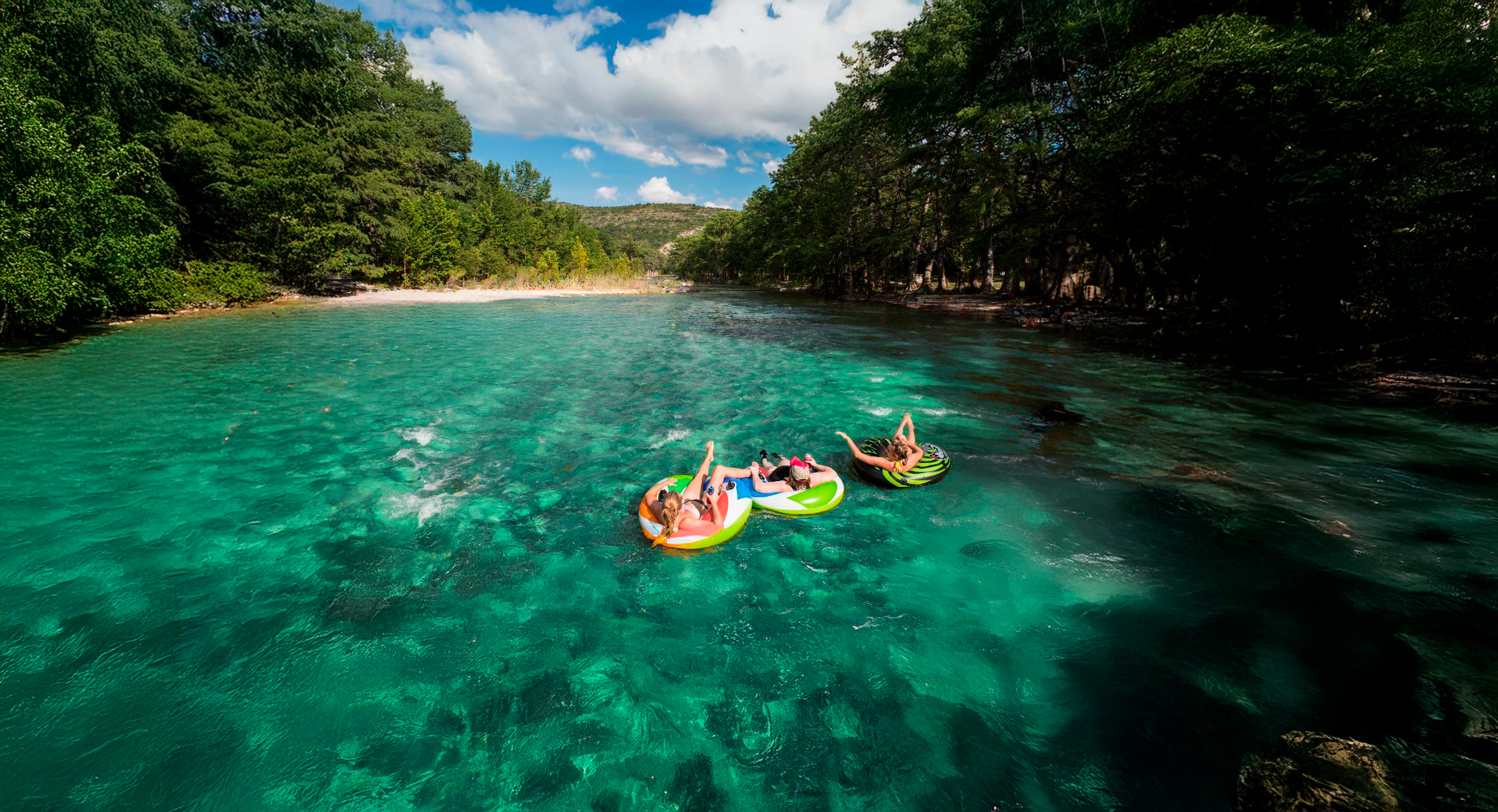 Three people are floating on colorful inflatable rafts in a crystal-clear river surrounded by lush green trees and rocky cliffs in Uvalde County, Texas