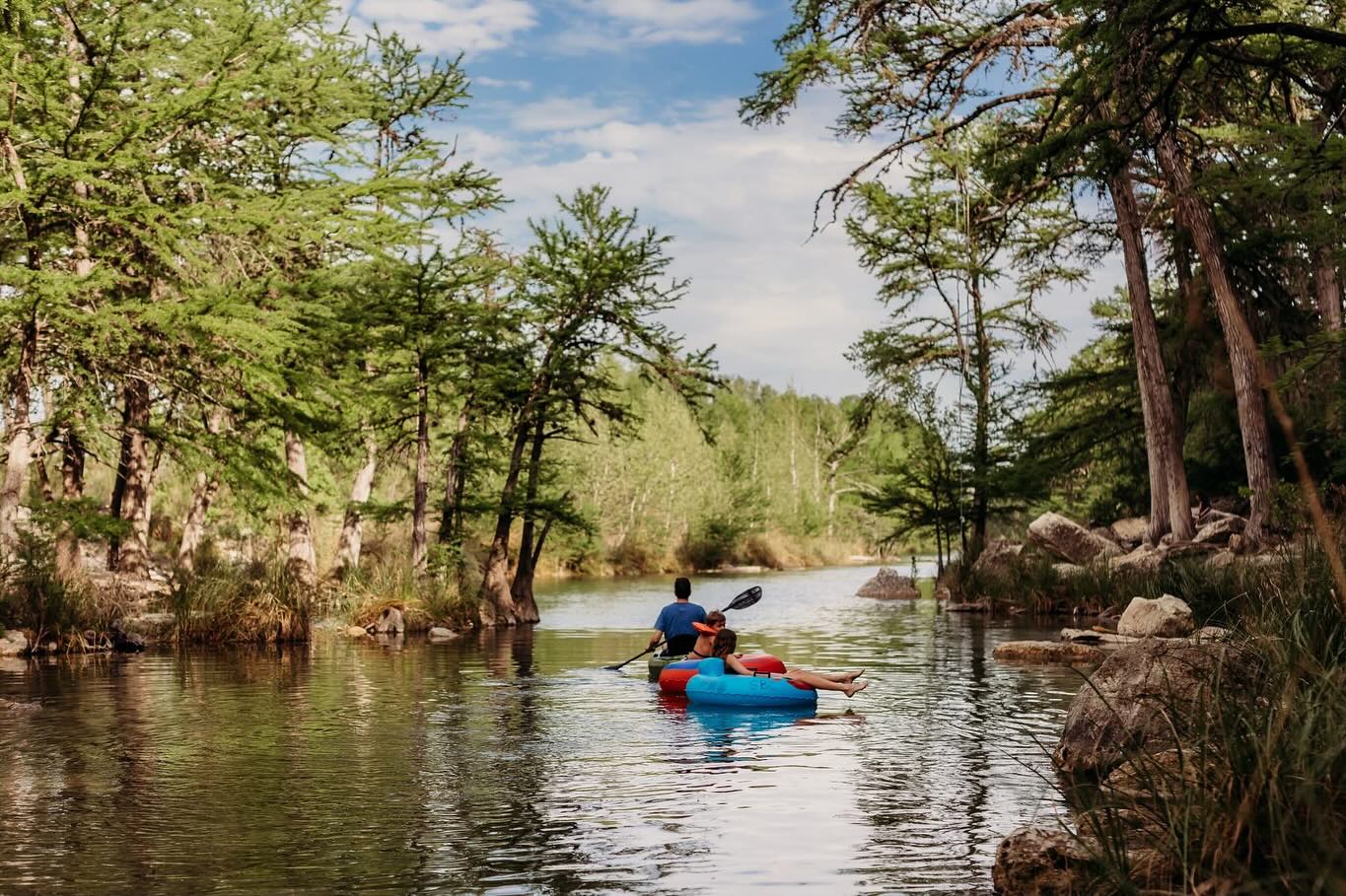 Two kayakers paddle down a serene river surrounded by lush green trees and rocks in Uvalde County, Texas