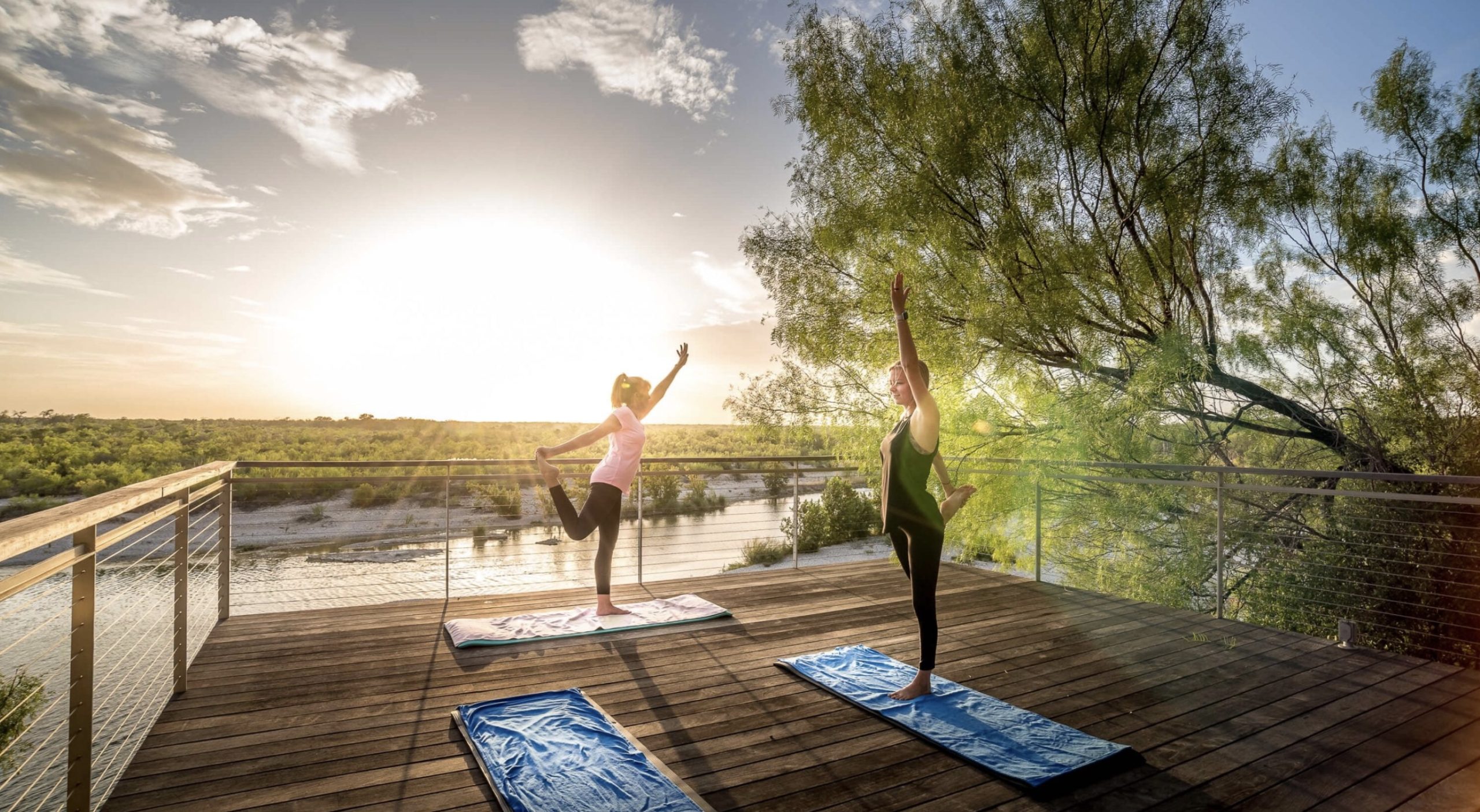 Two women practice yoga on a wooden deck overlooking a serene landscape at sunset in Uvalde County, Texas