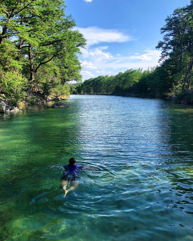 A person swims in a crystal-clear river surrounded by lush green trees and a bright blue sky in Uvalde County, Texas
