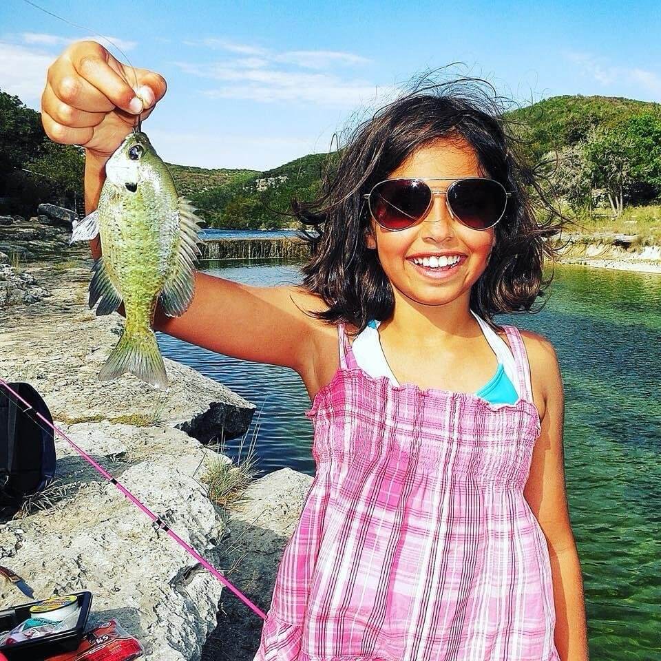 A young girl proudly displays her first fish catch, a small green fish, by a serene lake surrounded by lush greenery in Uvalde County, Texas