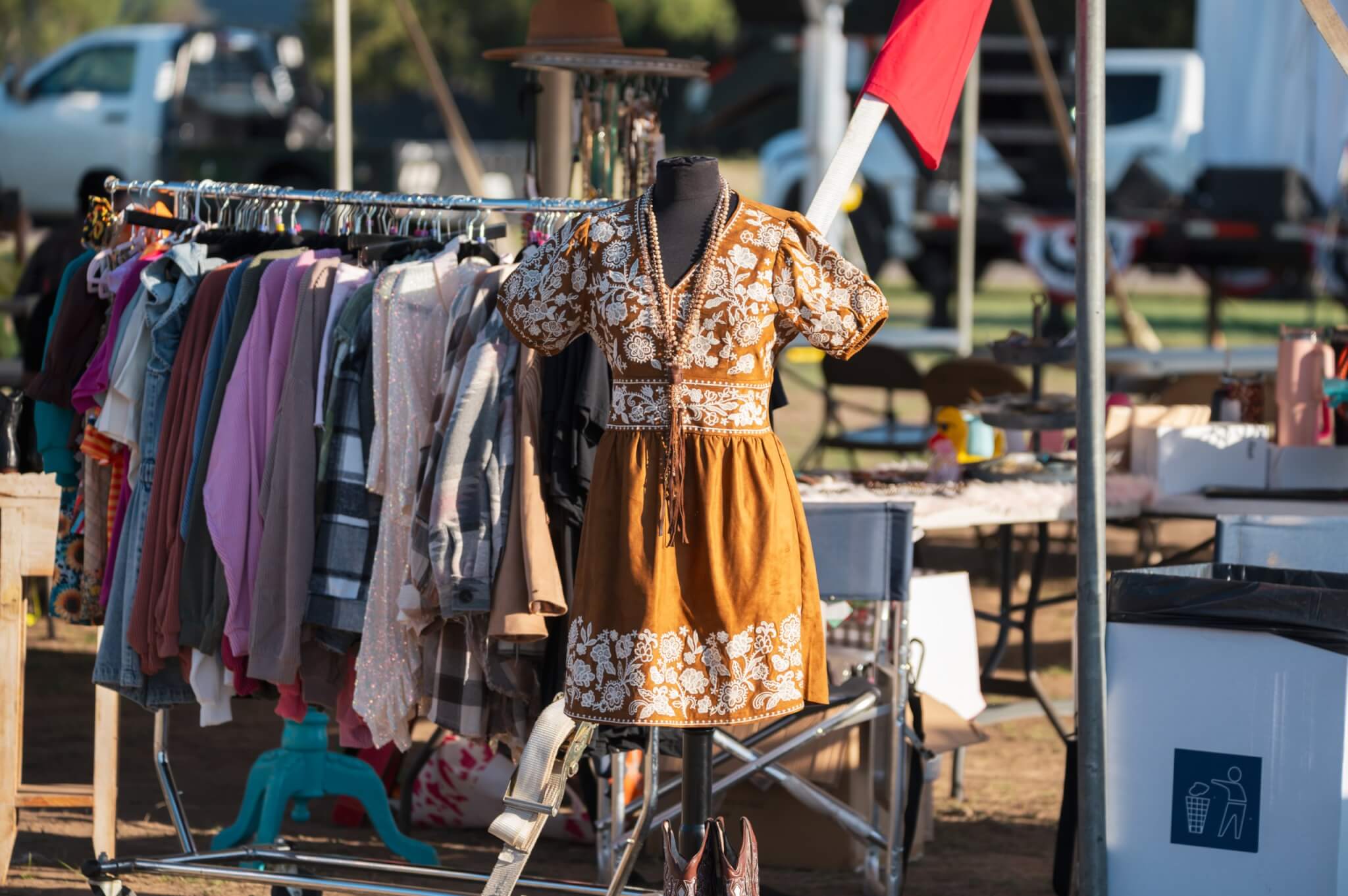 A mannequin dressed in a brown and white embroidered dress stands among racks of colorful clothing at an outdoor market in Uvalde County, Texas