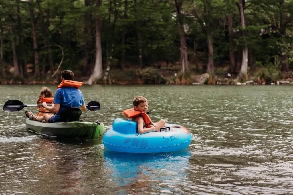 A family enjoys a leisurely day on the water, with two children in inflatable tubes and an adult paddling a kayak, all wearing life jackets in Uvalde County, Texas