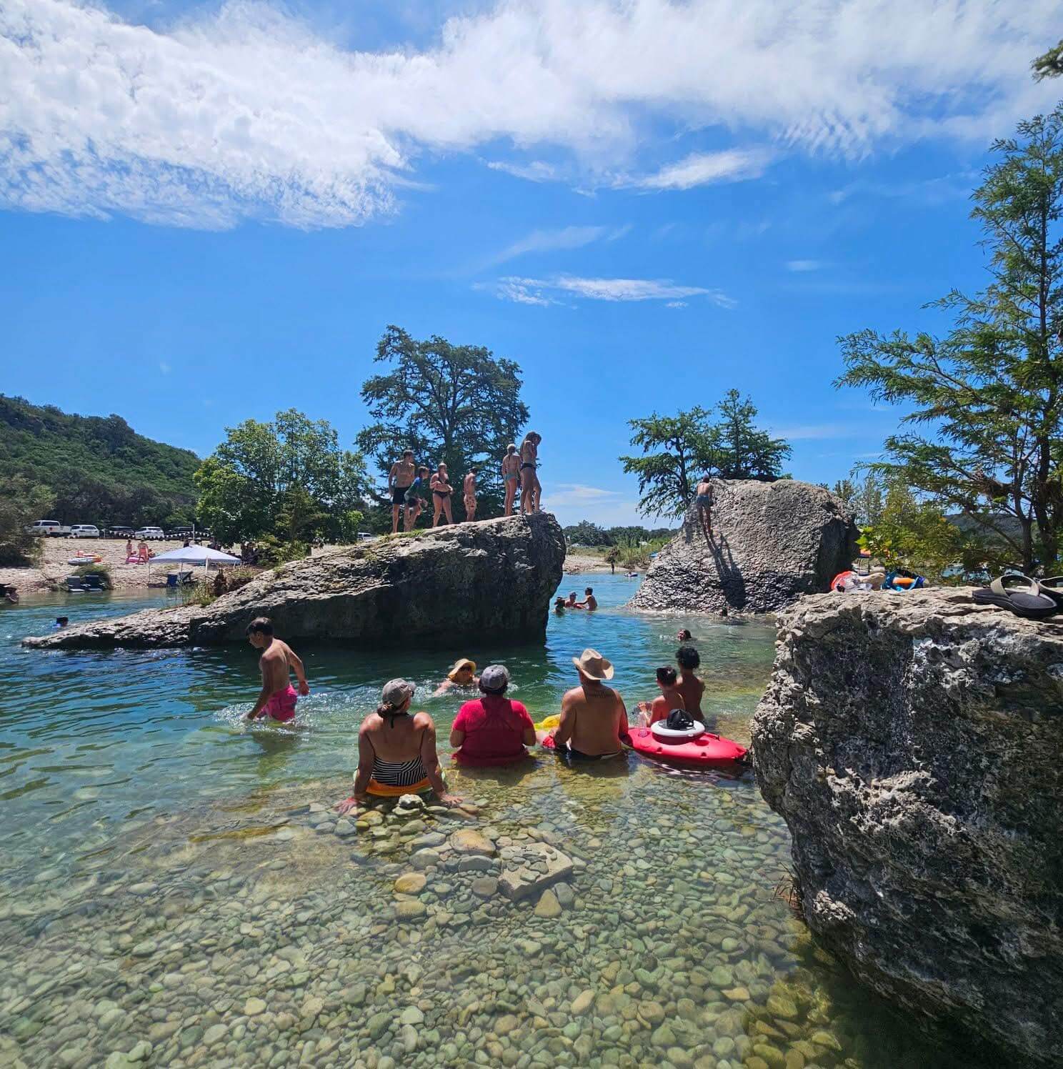 People are enjoying a sunny day at a clear, rocky river with some swimming and others relaxing on the rocks in Uvalde County, Texas