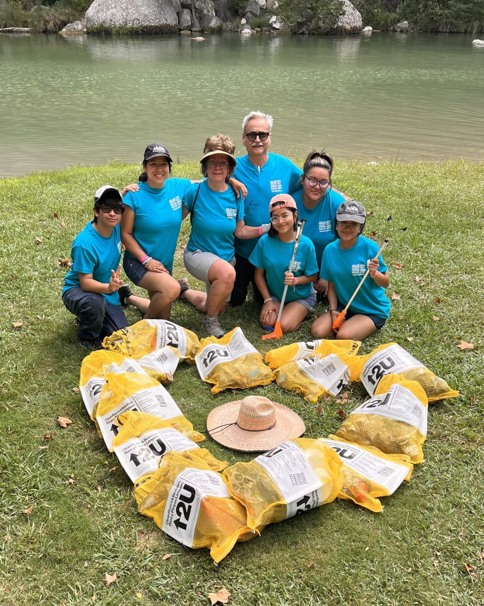 A group of eight people wearing blue shirts and hats are gathered around a large heart-shaped arrangement of yellow bags filled with leaves on a grassy area near a lake in Uvalde County, Texas