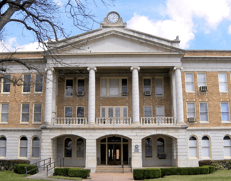 A grand, historic courthouse with a prominent clock tower and classical architecture stands majestically in a small town square in Uvalde County, Texas