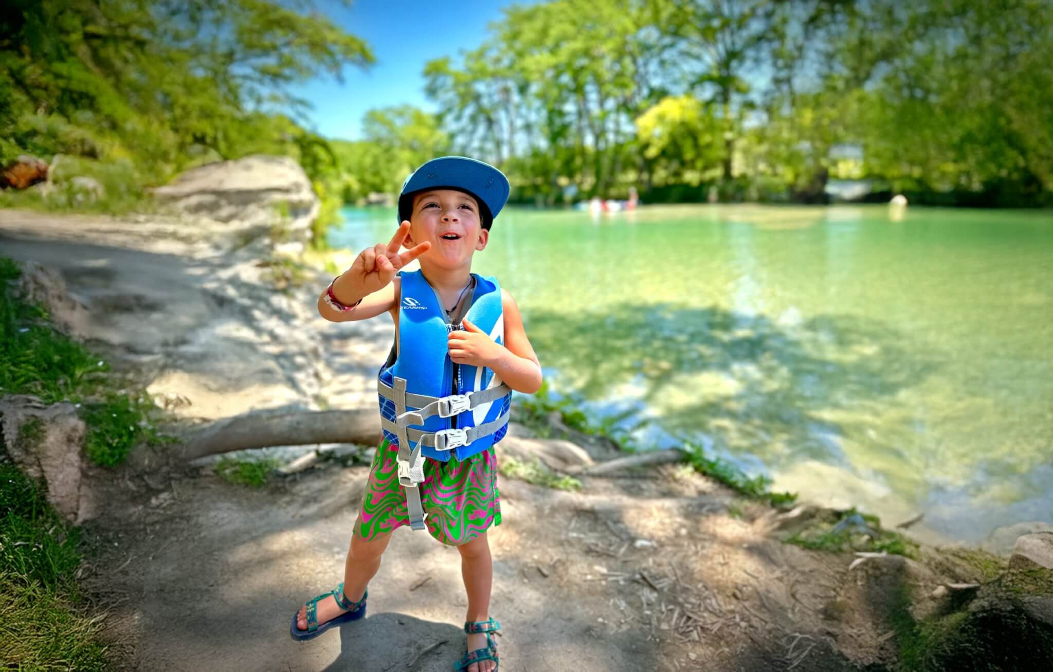 A young boy wearing a blue life jacket and hat stands on a dirt path near a serene, tree-lined lake, making a peace sign with his hand in Uvalde County, Texas