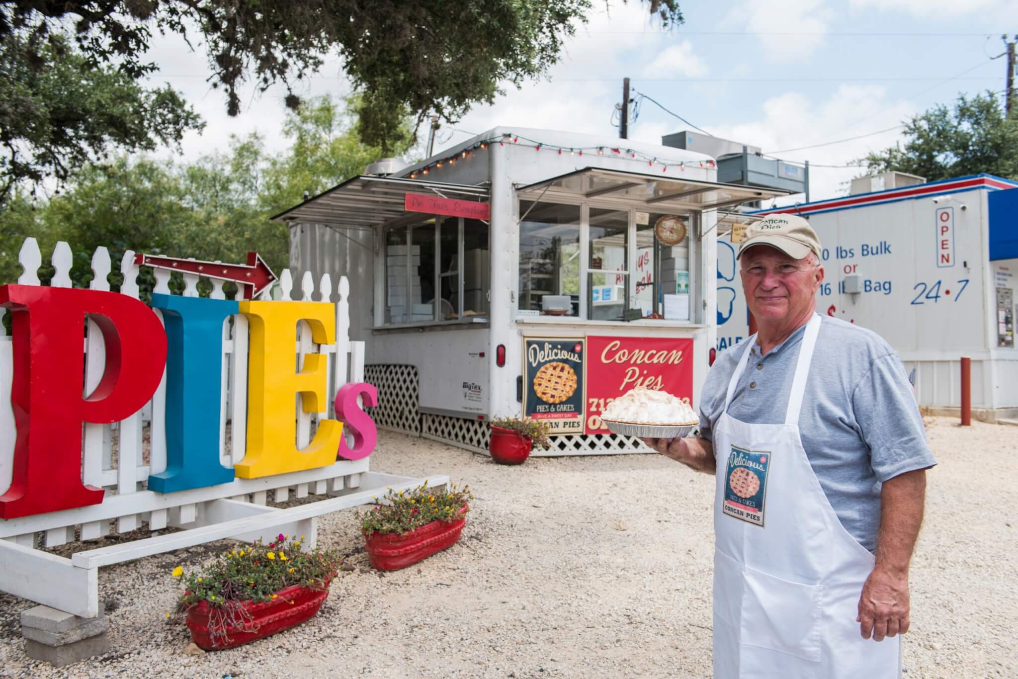 A man in a white apron stands outside a colorful pie shop, holding a pie, with a sign reading 'PIES' in large, vibrant letters in Uvalde County, Texas