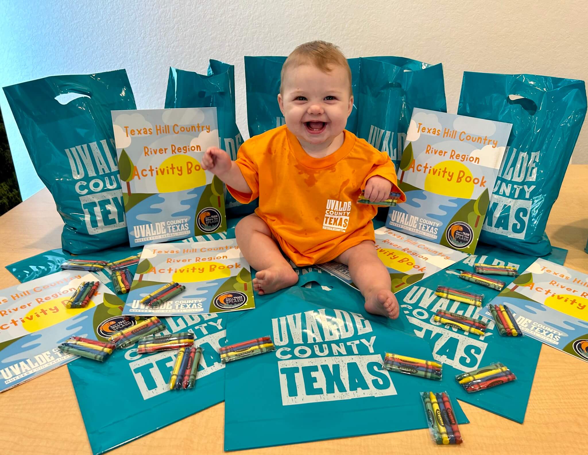 A cheerful baby sits amidst a colorful array of bags and activity books promoting Uvalde County, Texas, wearing an orange shirt that matches the vibrant theme in Uvalde County, Texas