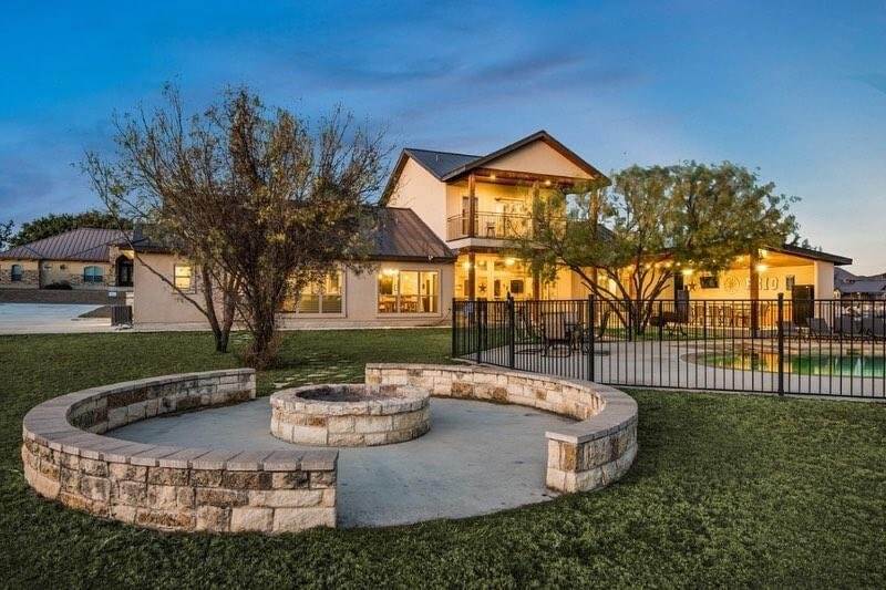 A spacious backyard features a circular stone fire pit surrounded by seating, a fenced swimming pool, and a large two-story house with illuminated windows and balconies in Uvalde County, Texas