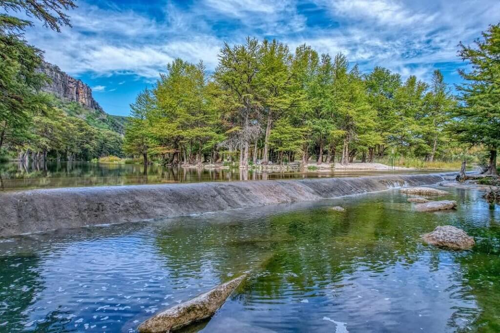Offers 1 A serene river winds through a lush, green forest with towering trees and rocky outcrops, reflecting the vibrant blue sky above in Uvalde County, Texas