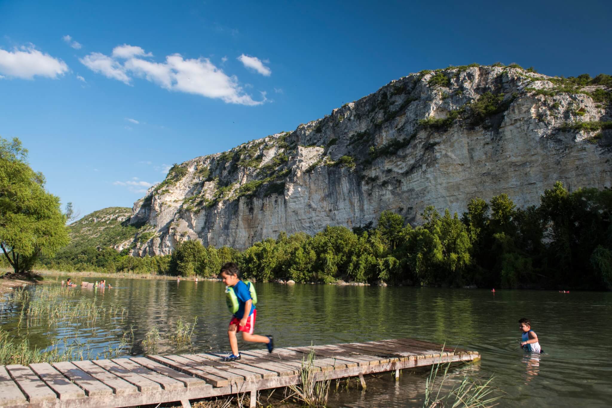 Lodging 21 A woman in a black bikini and sunglasses paddles a turquoise stand-up paddleboard on a serene river, accompanied by two small dogs wearing red life jackets in Uvalde County, Texas
