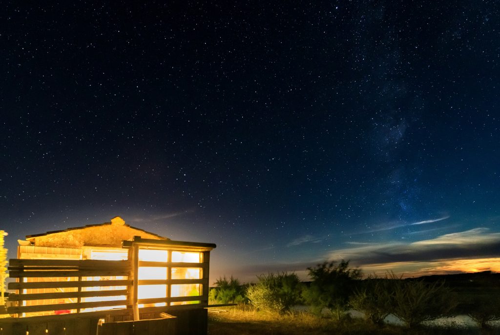 A rustic cabin with a glowing porch light stands alone in a vast, starry desert landscape under the Milky Way in Uvalde County, Texas