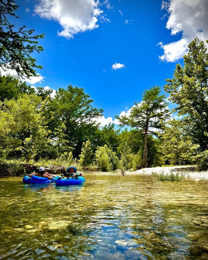 Two kayakers paddle down a serene, tree-lined river under a bright blue sky with scattered clouds in Uvalde County, Texas