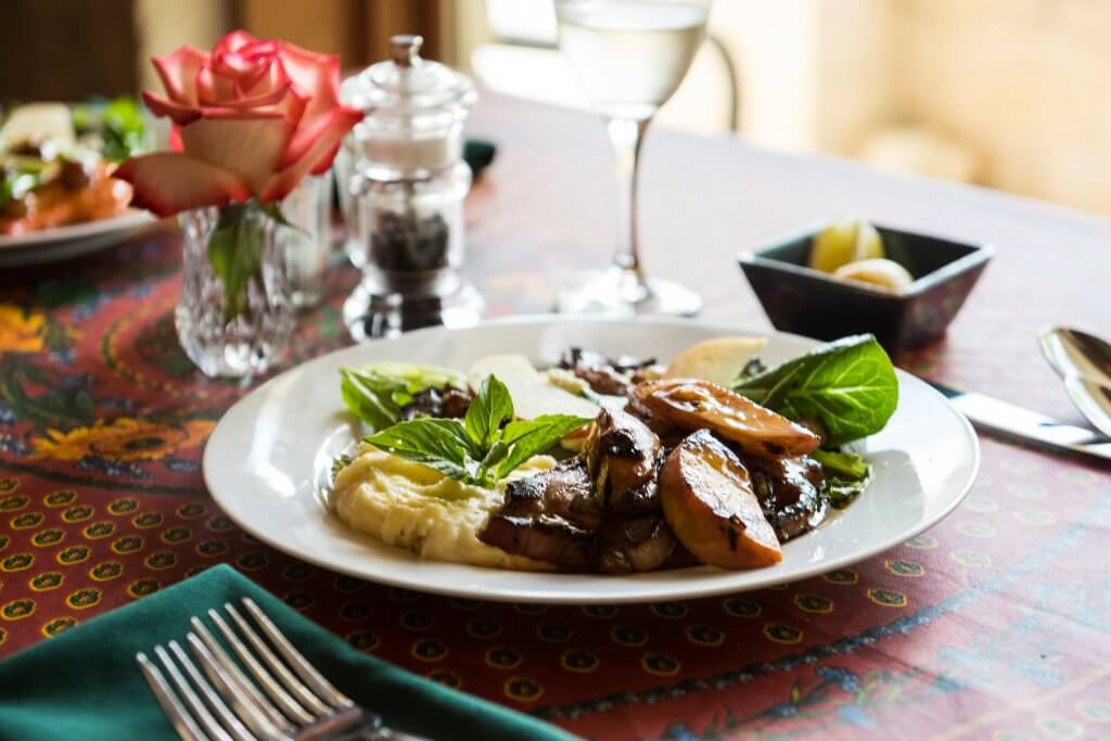 A beautifully presented plate of food featuring grilled mushrooms, mashed potatoes, and greens garnished with fresh herbs, accompanied by a glass of white wine and a rose on a colorful tablecloth in Uvalde County, Texas