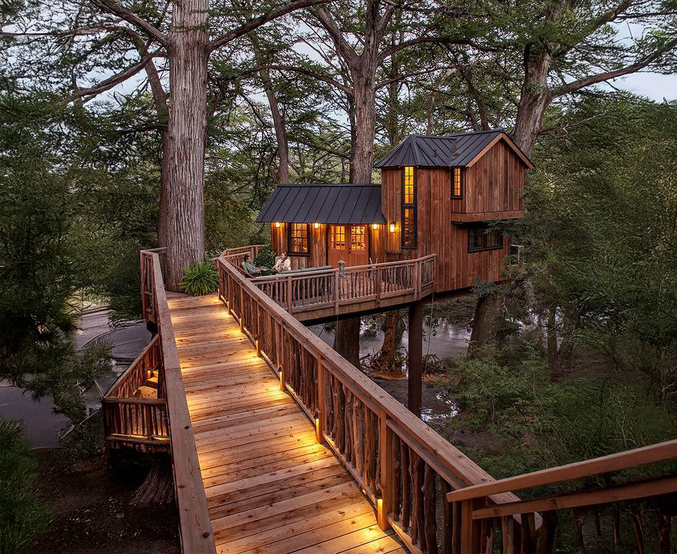 Lodging 2 A charming wooden treehouse nestled among towering trees, accessible by a winding wooden walkway with warm, inviting lights in Uvalde County, Texas