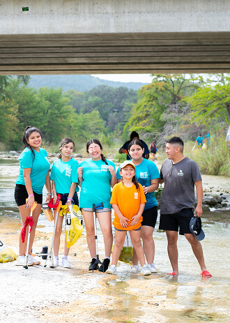 A group of people wearing blue shirts stand together under a bridge, holding cleaning supplies, ready to participate in a community service project in Uvalde County, Texas
