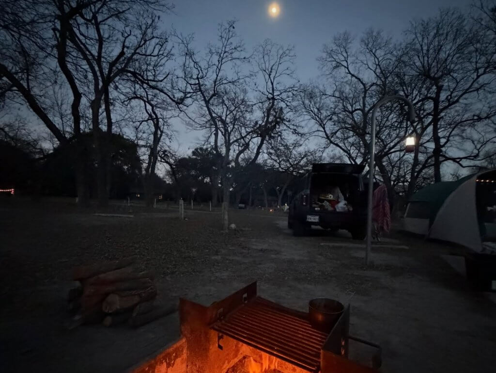 A serene night scene at a campsite with a crackling fire, illuminated by the soft glow of a full moon peeking through bare tree branches in Uvalde County, Texas
