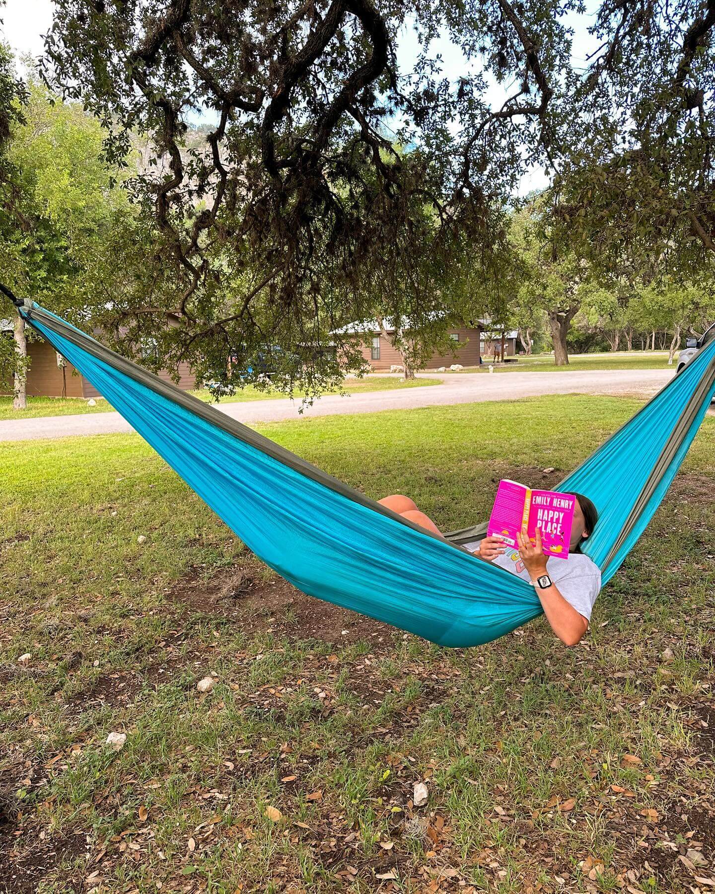 A person relaxes in a blue hammock under a tree, reading a pink book in a serene outdoor setting in Uvalde County, Texas