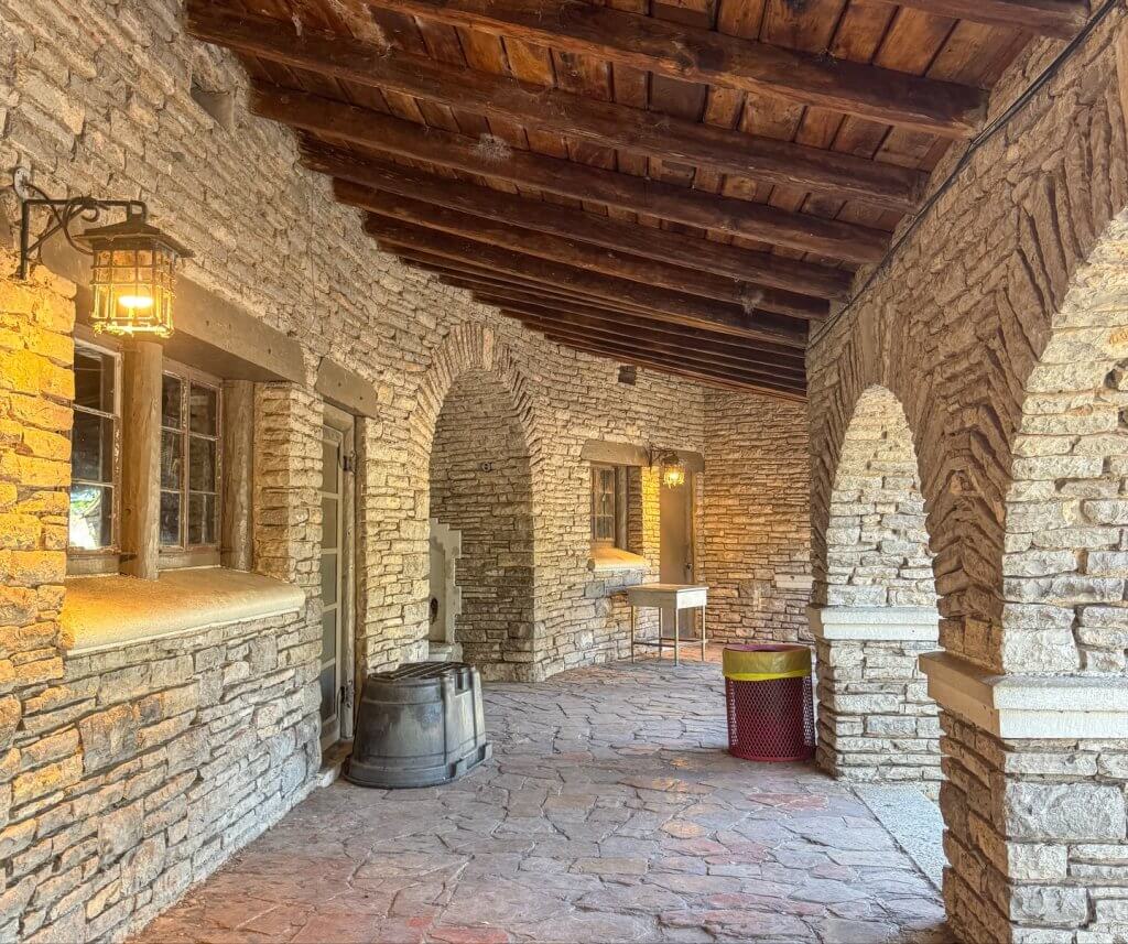 An arched stone corridor with wooden beams and lanterns, leading to a courtyard with a table and chairs in Uvalde County, Texas