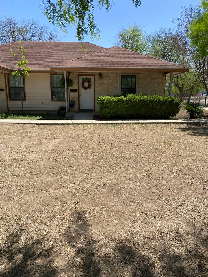 A single-story house with a brown roof and white siding is situated in a dry, grassy area with a few trees and bushes around it in Uvalde County, Texas