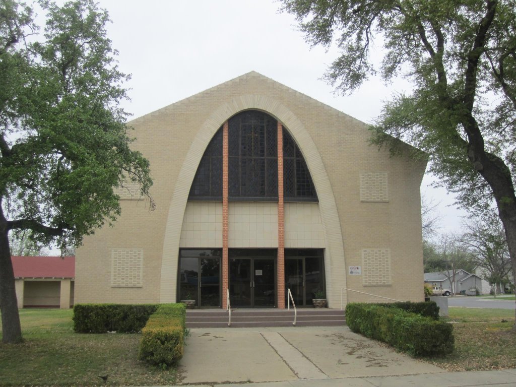 A modern church building with a distinctive curved facade and large arched windows, surrounded by greenery and trees in Uvalde County, Texas