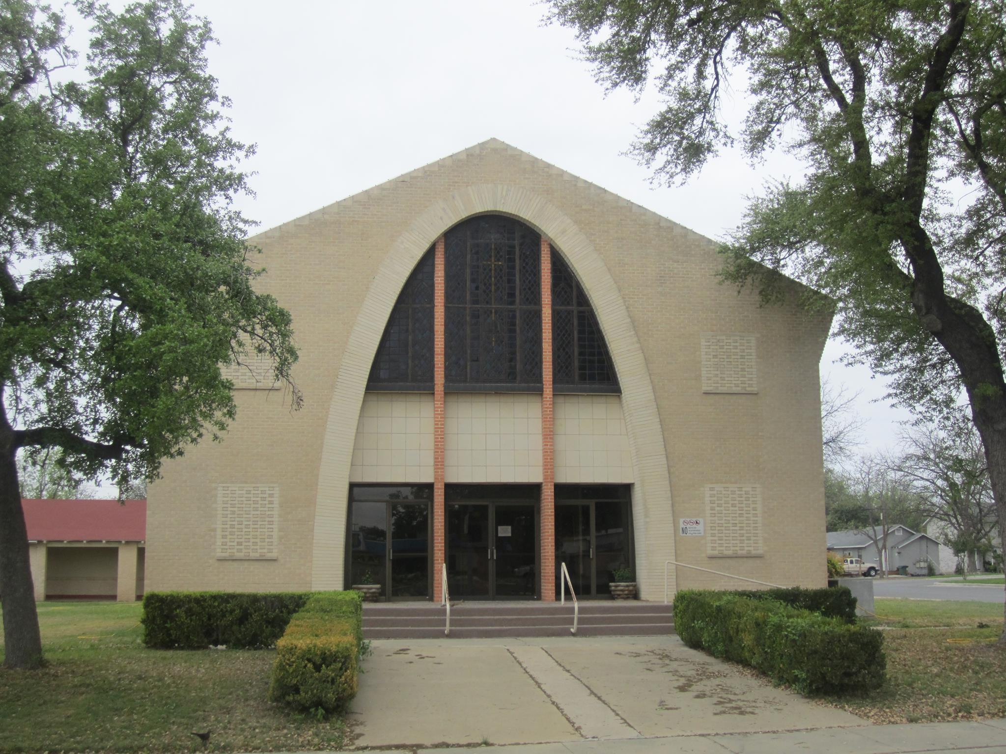 A modern church building with a distinctive curved facade and large arched windows, surrounded by greenery and trees in Uvalde County, Texas