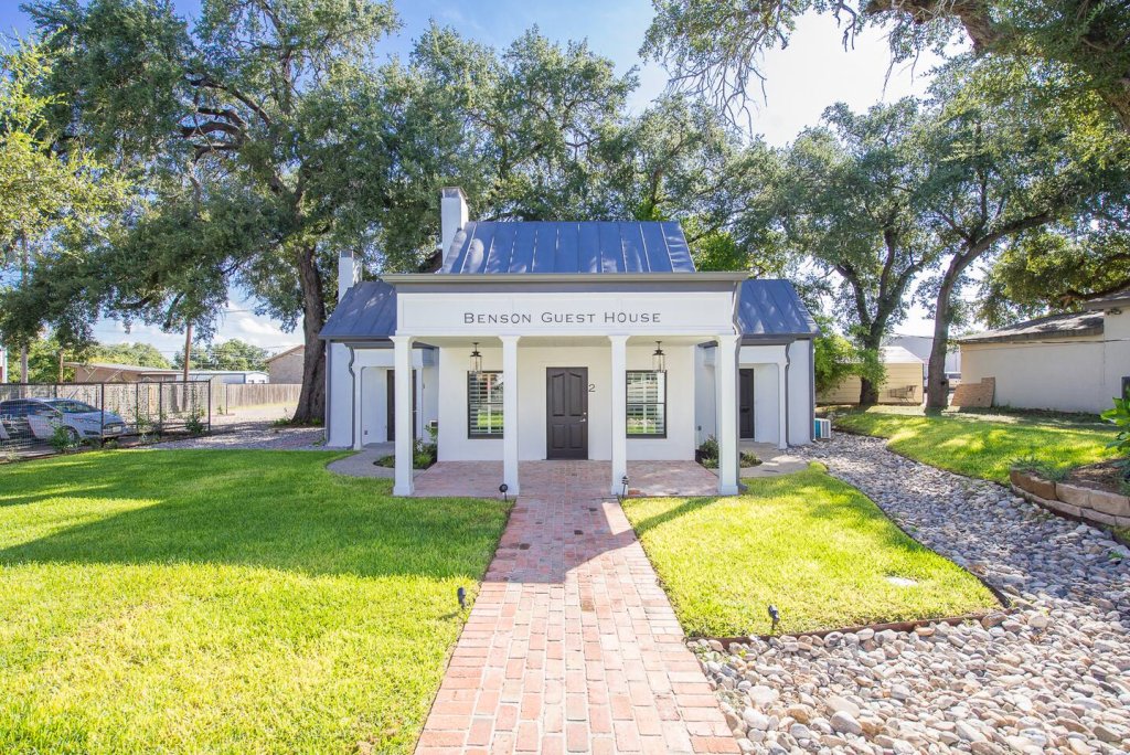 A charming white guest house with a blue roof, nestled among lush green trees and surrounded by a well-manicured lawn in Uvalde County, Texas
