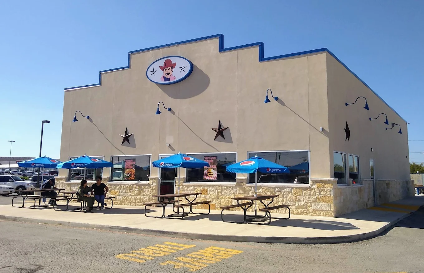 A bright, inviting restaurant with a cowboy theme, featuring outdoor seating under blue umbrellas and a large sign with a smiling cowboy mascot in Uvalde County, Texas