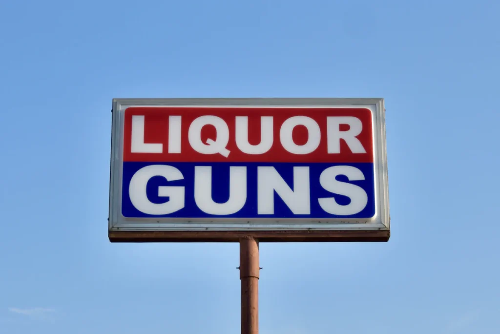 A large billboard sign with the words 'LIQUOR GUNS' in bold red and blue letters against a white background, mounted on a pole against a clear blue sky in Uvalde County, Texas