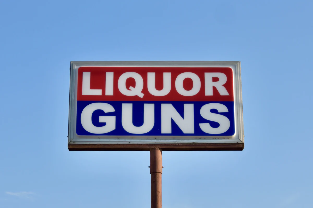 A large billboard sign with the words 'LIQUOR GUNS' in bold red and blue letters against a white background, mounted on a pole against a clear blue sky in Uvalde County, Texas