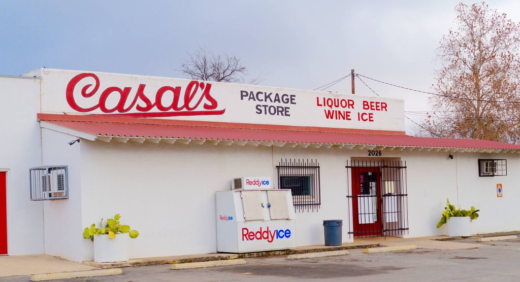 Casals Package Store, a white building with a red roof, features a large sign advertising liquor, beer, wine, and ice in Uvalde County, Texas