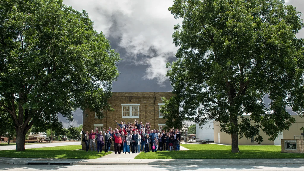 A large group of people, possibly a church choir, stands in front of a brick building surrounded by lush green trees and a clear blue sky in Uvalde County, Texas