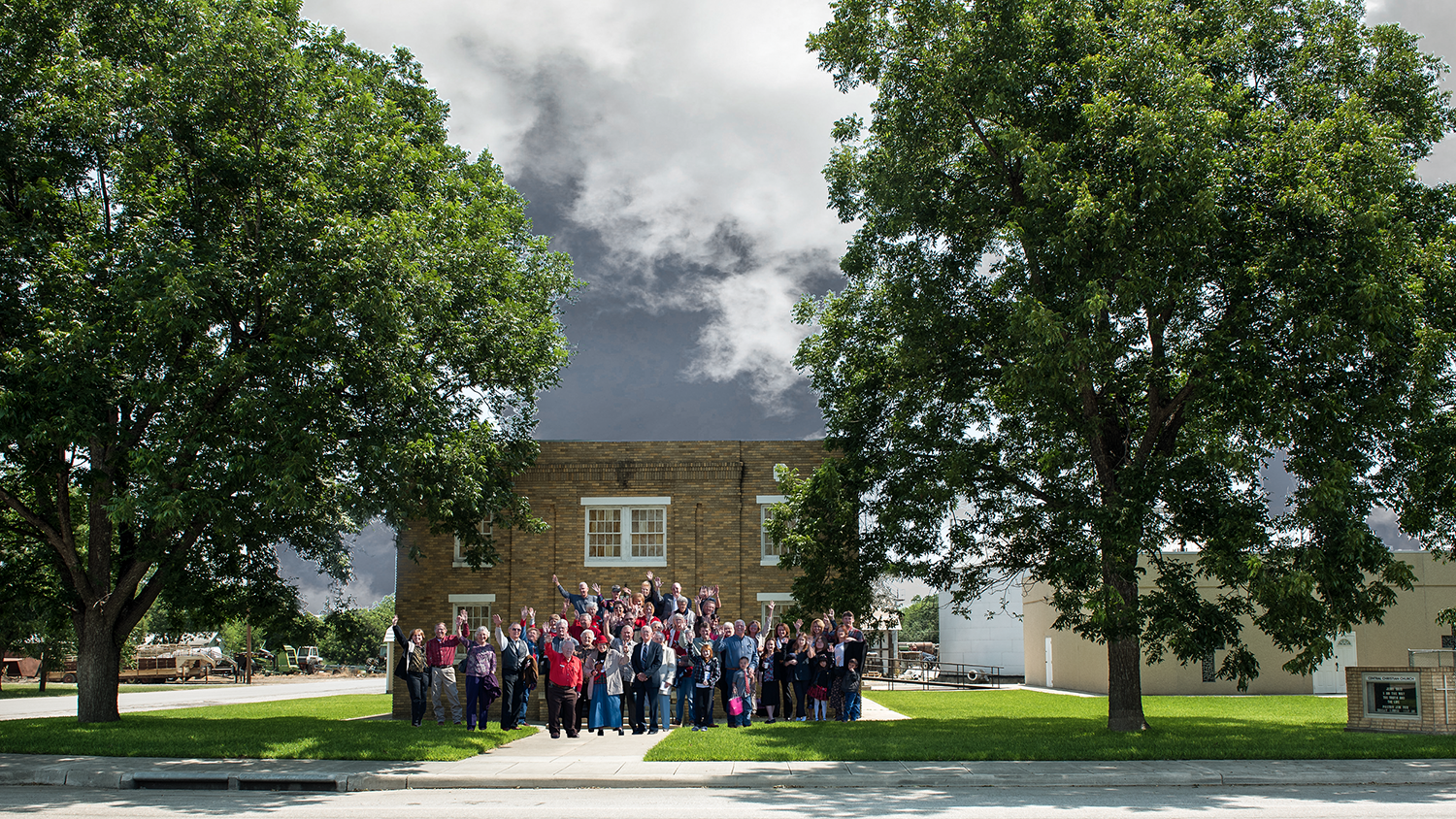 A large group of people, possibly a church choir, stands in front of a brick building surrounded by lush green trees and a clear blue sky in Uvalde County, Texas