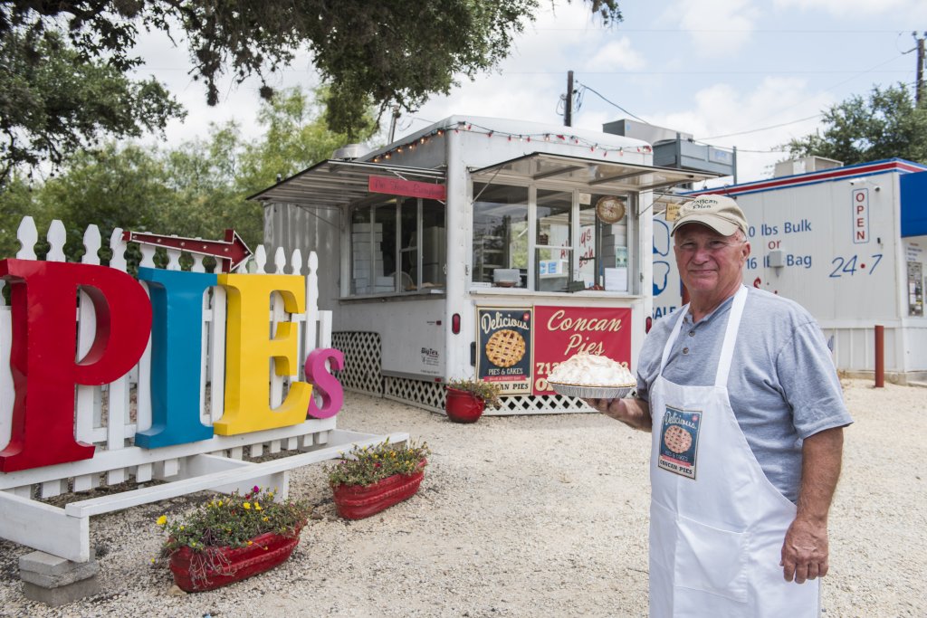 A man in a white apron stands in front of a colorful pie stand with large, painted letters spelling 'PIES' in bright colors in Uvalde County, Texas