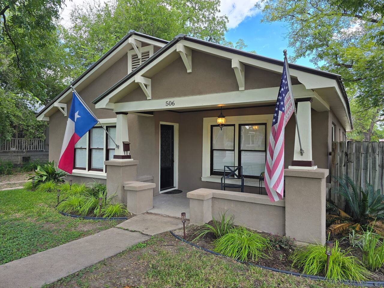 A charming two-story house with a covered front porch, adorned with American and Texas flags, set against a backdrop of lush green trees in Uvalde County, Texas