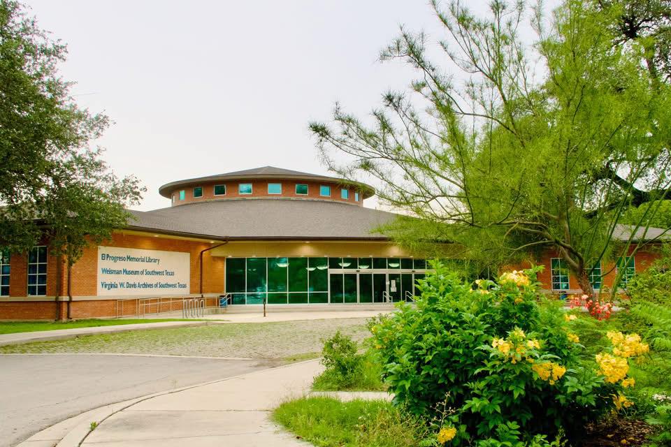 A modern, circular building with a green roof and large glass windows is nestled among lush greenery and blooming yellow flowers in Uvalde County, Texas