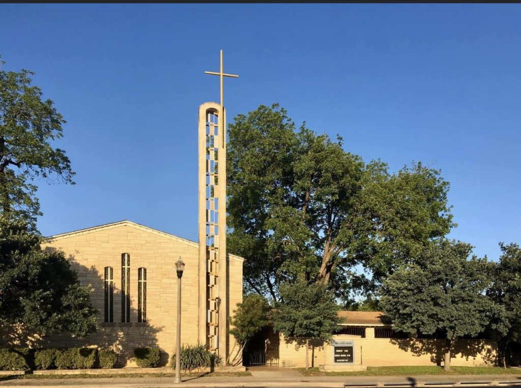 A church with a tall bell tower and a cross on top, surrounded by lush green trees under a clear blue sky in Uvalde County, Texas