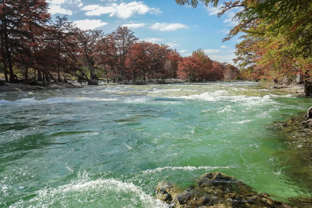 A rushing river winds through a vibrant autumn forest, with clear turquoise waters contrasting against red and orange trees in Uvalde County, Texas