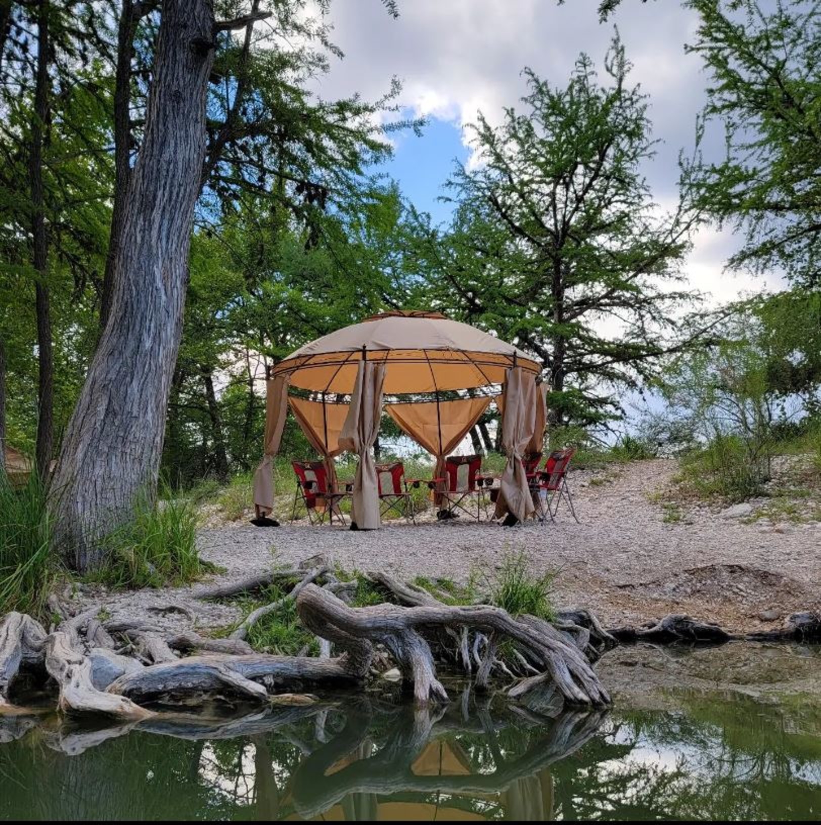 A serene outdoor setting with a beige canopy tent surrounded by lush green trees and a tranquil pond in Uvalde County, Texas