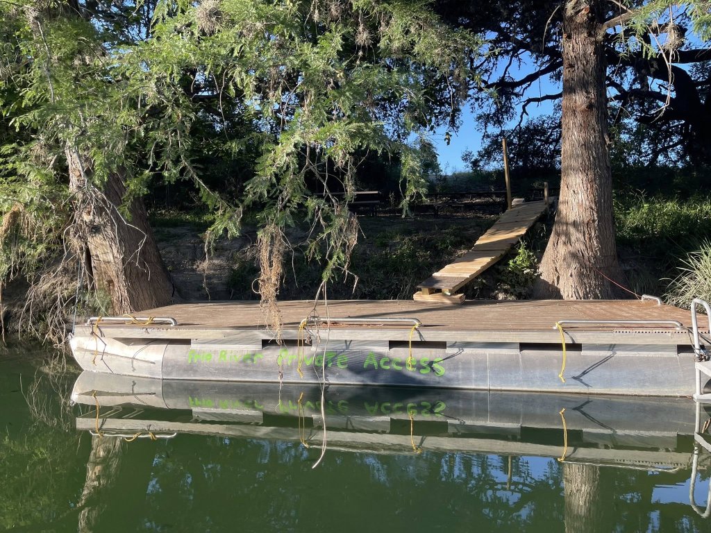 A serene scene featuring a wooden dock extending over a calm, green body of water, surrounded by lush trees and vegetation in Uvalde County, Texas
