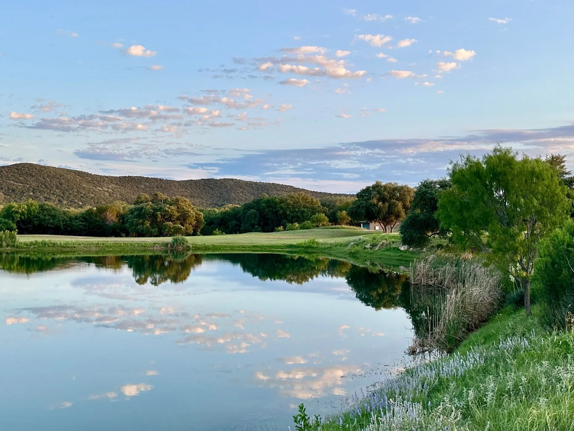 A serene lake reflects the surrounding trees and distant hills under a clear blue sky with scattered clouds in Uvalde County, Texas