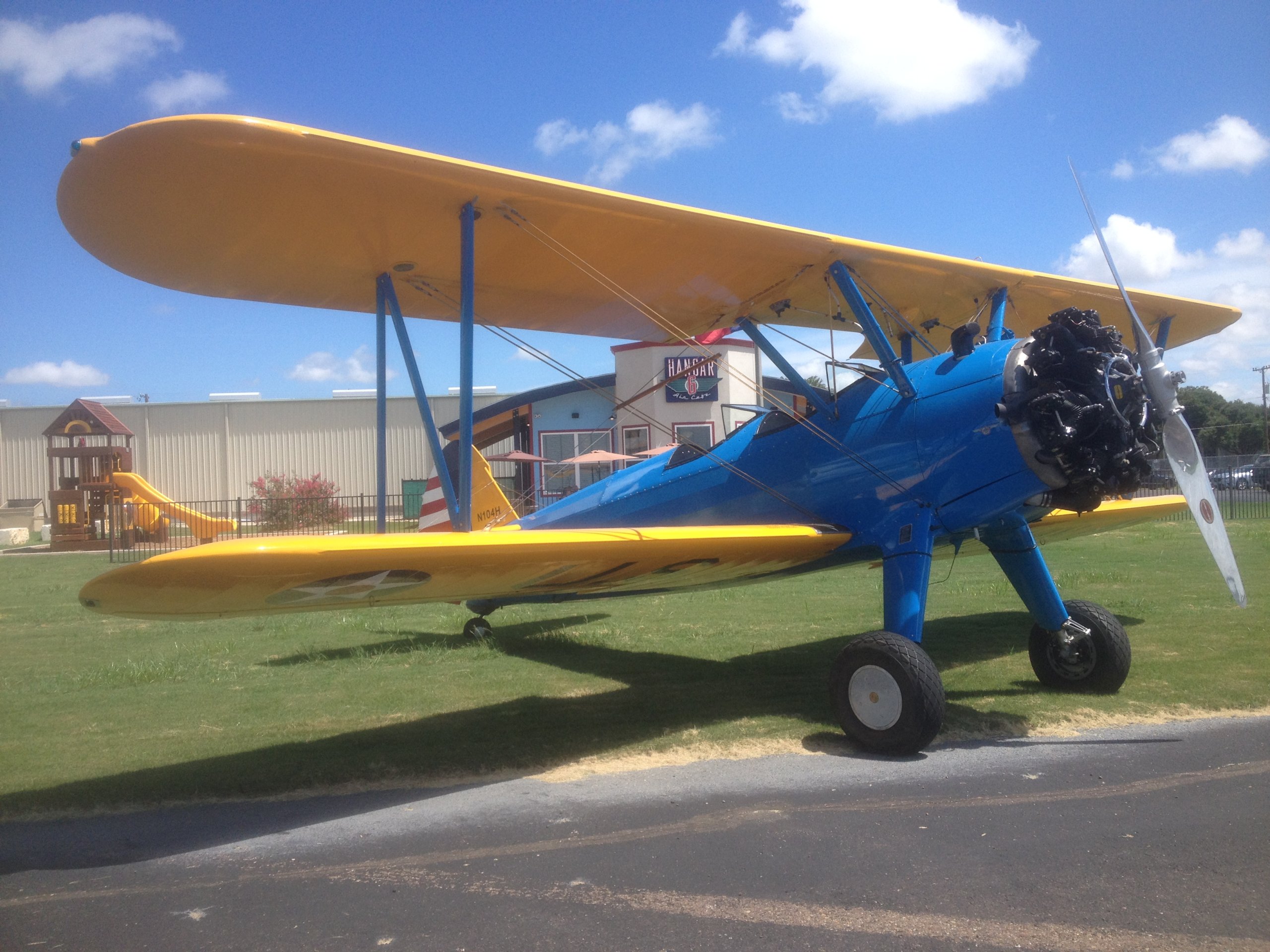 A vintage biplane with yellow wings and a blue fuselage is parked on a grassy field under a clear blue sky in Uvalde County, Texas