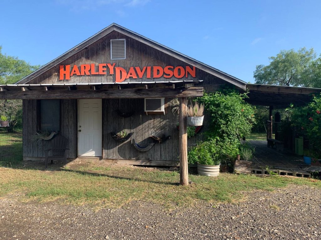A rustic Harley-Davidson dealership with a wooden exterior and a prominent sign, surrounded by greenery and a gravel parking area in Uvalde County, Texas