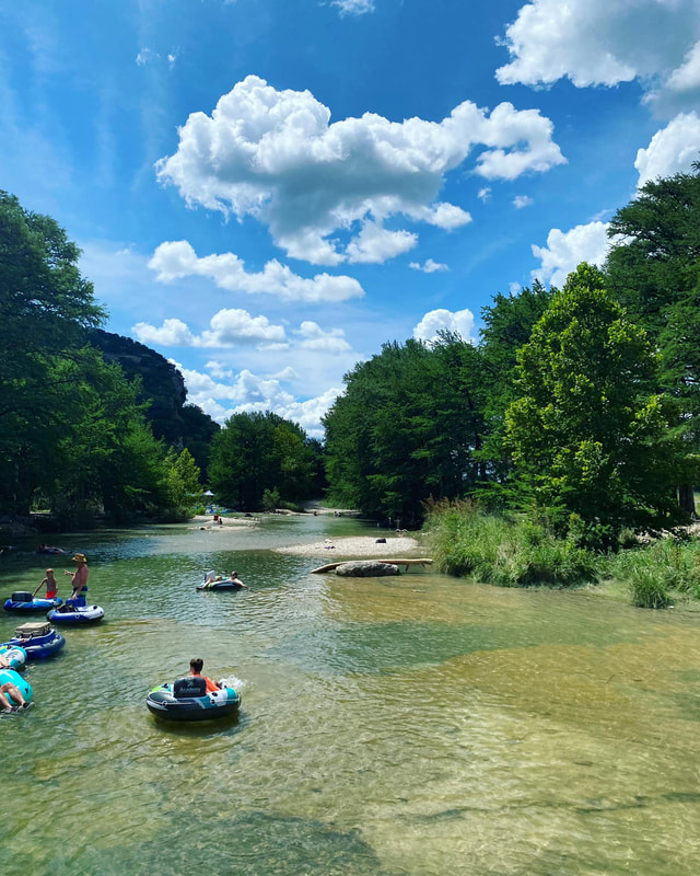 A serene river winds through a lush, green forest, dotted with people enjoying a sunny day on inflatable tubes in Uvalde County, Texas