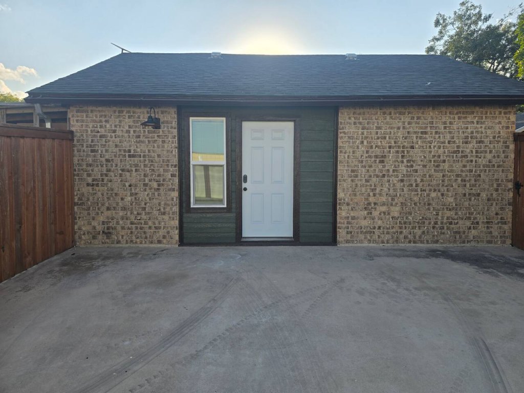 A small, single-story brick house with a white front door and a single window, set against a backdrop of a clear blue sky and surrounded by a wooden fence in Uvalde County, Texas
